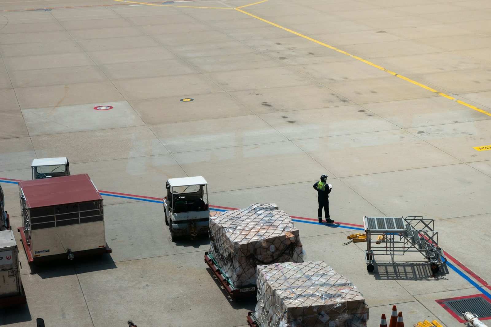 Wrapped cargo pallets and tugs on an airport apron beside a ground marshal