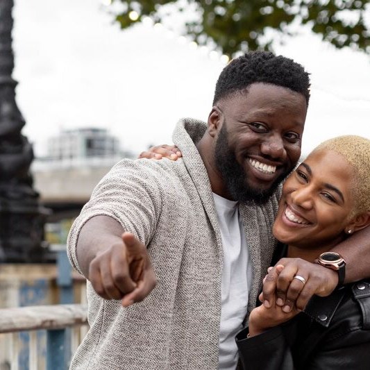 A smiling couple pointing ahead while a friend photographs them on a city waterfront